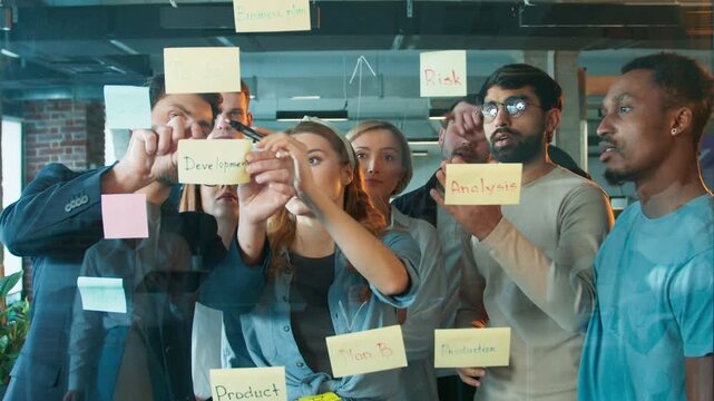 Group of people organizing sticky notes on glass. Team reviewing tasks while arranging labels like development and analysis. Focused woman observing layout as colleagues adjust strategy.