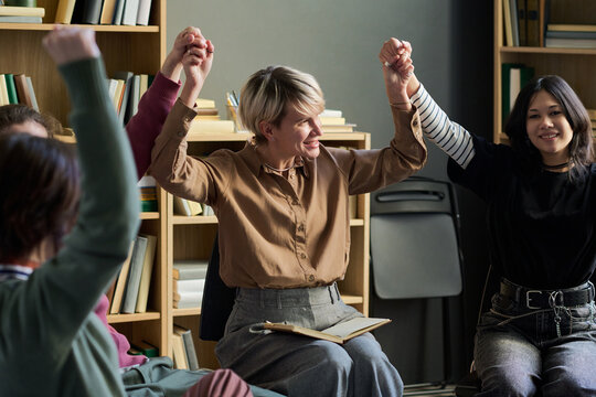 Diverse group of adult and young women and men sitting in circle holding hands raised during group therapy session, smiling and in supportive activity together