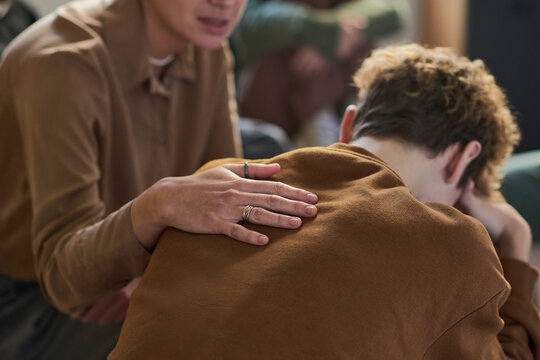 Teenager sitting with head in hands being comforted by young Asian man during group therapy session, supportive gesture showing empathy and emotional support - Powered by Adobe