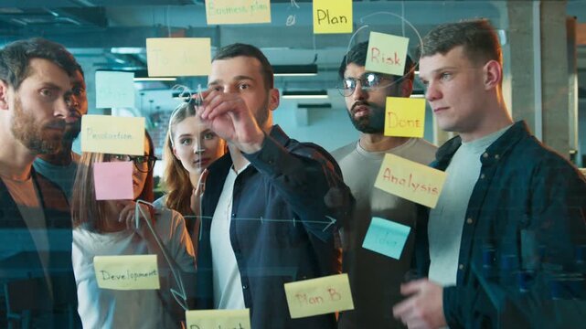 Handsome male guiding team while drawing workflow on glass board. Focused group studying sticky notes closely. Team planning new project steps together in bright office environment.