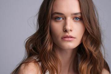 Retrato elegante de mujer joven con cabello casta&ntilde;o ondulado y ojos azules, posando en estudio con iluminaci&oacute;n suave y expresi&oacute;n serena.