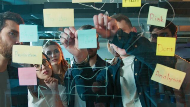 Team of coworkers arranging idea note on glass board. People focusing on workflow layout. Group shaping project structure during planning session inside new office location. Helping each other.