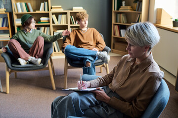 Young woman writing on clipboard while two young adults, one man and one woman, sitting in chairs talking during group therapy session in library setting