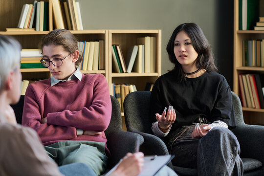 Diverse group of young people and teenagers participating in group therapy session, teenage boy sitting with arms crossed while Asian young woman speaking and gesturing to therapist