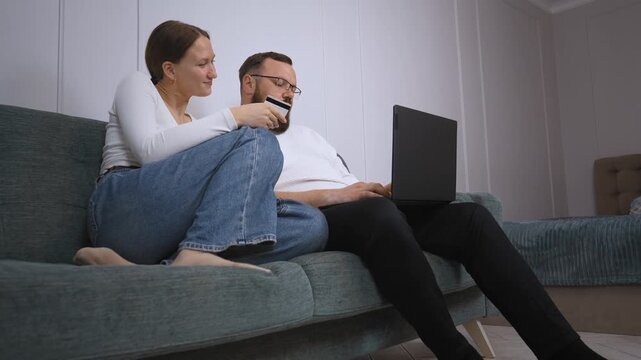 Young couple sitting on couch and using world wide web for home shopping and using credit card and laptop to shop online