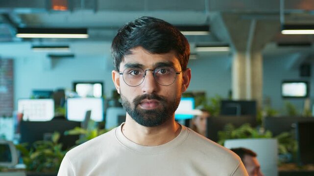 Serious male staring directly at camera. Bearded man standing still with focused expression. Professional worker posing for portrait shot. In office space with monitors and plants behind.
