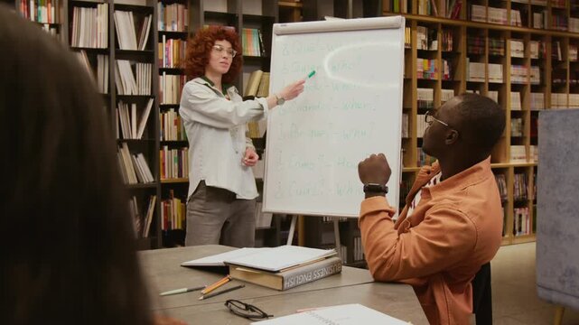 Young female Spanish teacher presenting special question words on whiteboard to adult students sitting at library table in small study group for beginners