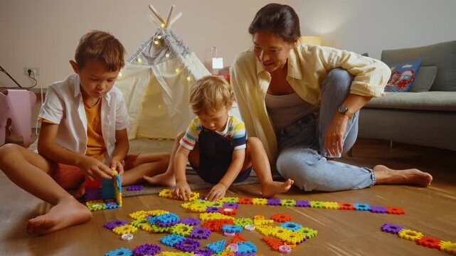 Young smiling mother and her two sons playing with a colorful constructor puzzle on the floor in the living room, enjoying a creative and educational activity together on a family weekend