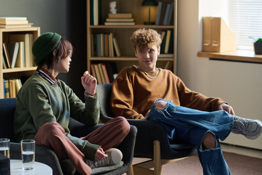 Two teenagers sitting in chairs in conversation during group therapy session, one teenager listening attentively while other speaking, bookshelves in background