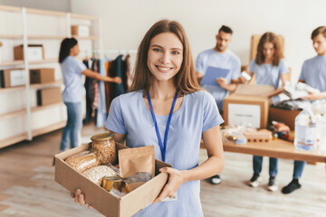 A group of volunteers collects and sorts food donations in a community center. One young woman smiles while holding a box filled with food items. Others are busy preparing donations.