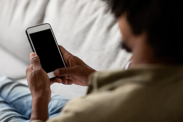 Black male sits on a couch in his living room, casually browsing the internet on a blank smartphone. The atmosphere is relaxed and comfortable, emphasizing leisure time at home.