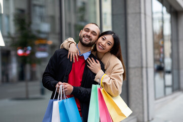 A joyful couple stands outside a store on a bustling city street. They are smiling widely, holding...
