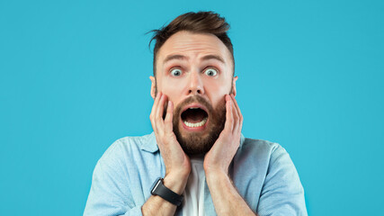 A man with a beard looks surprised as he holds his face with both hands. His eyes are wide open, and he seems to be reacting to something unexpected. The background is bright blue.