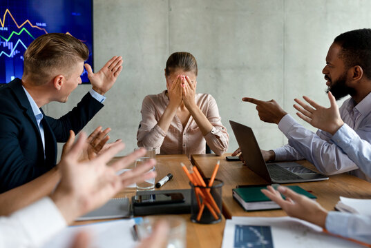 In a tense corporate meeting, a group of coworkers shouts at a young businesswoman, pointing fingers and expressing frustration. She appears overwhelmed and covers her face with her hands.