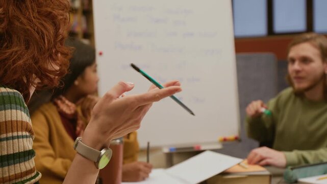 Close up on hand of curly red haired woman twirling pencil thinking over grammar rules while discussing new topic with male teacher in group English class in library