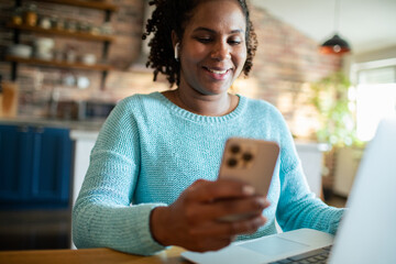 Adult woman smiling at smartphone while using laptop at home kitchen