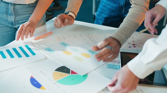Hands of colleagues leaning over table. Team reviewing printed charts and diagrams. Markers and tablet on surface. Group analyzing workflow steps in office room. Data sheets guiding discussion.