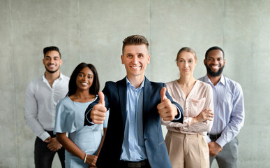 A team of diverse business people poses in an office against a grey wall. The male leader smiles and gives a thumbs up, promoting their company's services with confidence.
