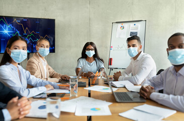 Diverse group of business professionals wearing protective masks have a meeting in an office. Colleagues look at the camera while discussing strategies during the pandemic.