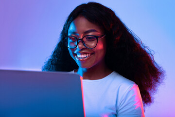 A young woman with curly hair is smiling while looking at her laptop screen. The bright, colorful lighting creates a vibrant atmosphere as she interacts with technology.