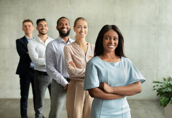 A confident black female leader stands with her multiethnic team in a stylish office. All team members smile and convey a spirit of collaboration and modern entrepreneurship.