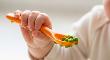 Close-up of child's hand holding orange spoon with green peas, symbolizing infant feeding, nutrition, or early childhood food introduction