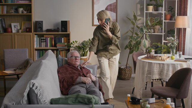 Long shot of stressed adult Caucasian woman calling doctor on mobile phone while taking care of sick senior father lying on couch with fever and shivering