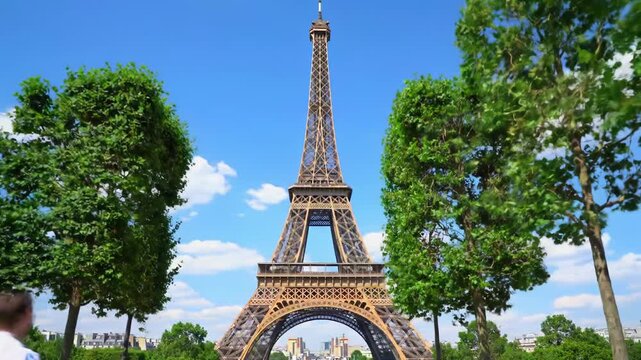 A slow wide-angle dolly out video of the majestic eiffel tower in paris, framed by lush green trees, with the sprawling cityscape under a clear blue sky, crisp natural daylight. Serene parisian
