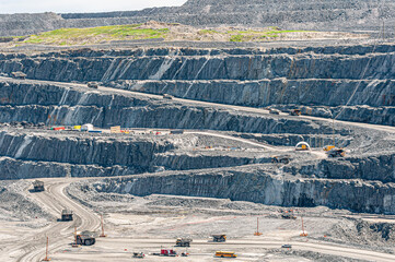 Large open-pit mine with terraced rock walls, heavy trucks, machinery and service structures. A detailed view of industrial extraction and quarry activity under bright daylight.
