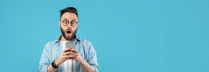 Naklejka premium A young man with glasses and a beard expresses shock while checking his smartphone. He stands against a bright blue background, highlighting his surprised reaction.