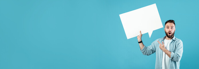 Fototapeta premium A young man with a surprised expression holds a large blank speech bubble. He stands against a solid blue backdrop, ready to convey a message. His gesture invites curiosity from viewers.