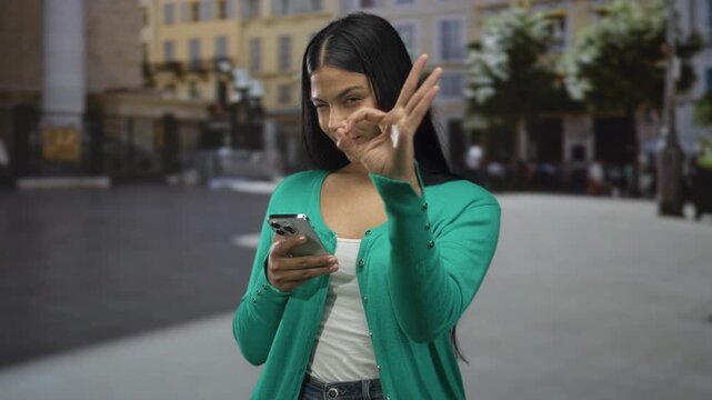 Young latin woman scrolls smartphone screen checking message alerts and makes ok sign on urban street corner; approval.
