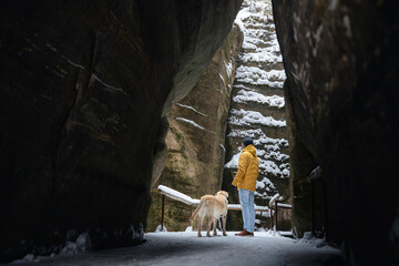 Man walks with dog along narrow snowy path between high rocks. Labrador retriever stays close to...