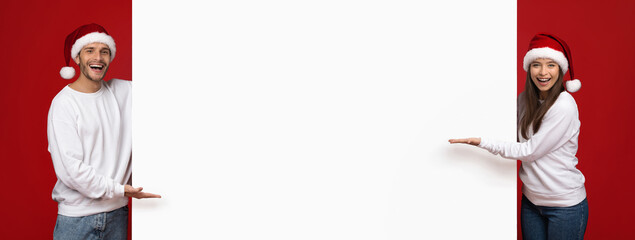 Cheerful couple wearing Santa hats stands on either side of an empty sign. They both smile...