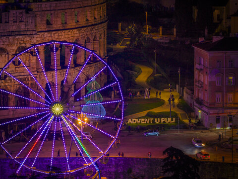 Aerial view of the brilliantly lit Ferris wheel stands in stark contrast to the ancient amphitheater, illuminating the "Advent u Puli" celebration, Pula, Croatia.