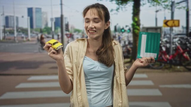 Young woman smiling, holding a yellow toy car and a learner plate at a city street crosswalk; learning independence confidence.