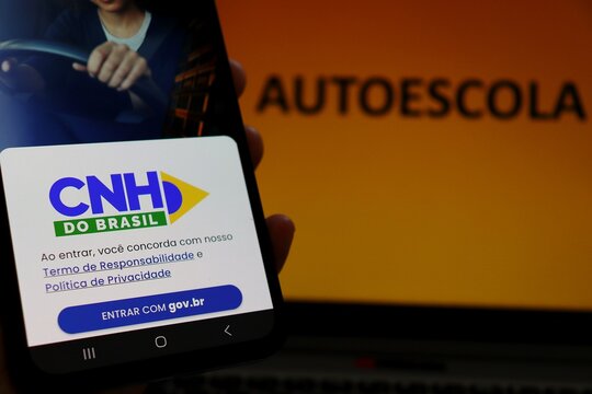 Salvador, Brazil - December 9, 2025: CNH Digital app on smartphone screen and "Autoescola" (driving school) sign in the background. Driving license process in Brazil. 