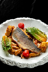 A gourmet dish of pan-seared sea bass with fresh baby gem lettuce, grilled corn, and cherry tomatoes on a white artistic plate against a black background