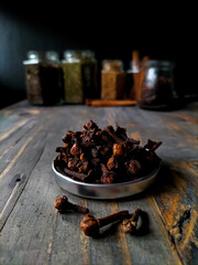 A small, silver bowl filled with dried cloves sits on a dark, rustic wooden surface. 