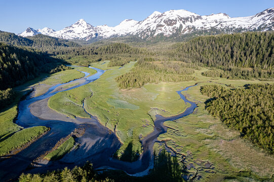 Aerial view of a meandering river carving through a verdant valley floor, bordered by dense forests and crowned by snow-capped peaks, Cordova, Alaska, United States.