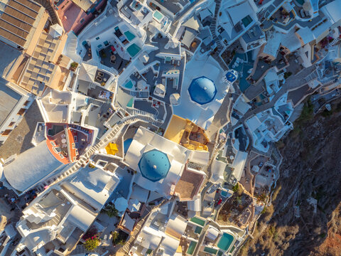Aerial view of white buildings cascading down the cliffside, punctuated by the vibrant blue domes of churches, contrasting with the rugged terrain, Oia, Thira, Greece.