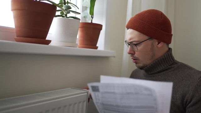 Worried man in a chilly apartment reacts with surprise to his soaring utility bills and heating costs, frantically inspecting a radiator while grappling with expenses amidst the energy crisis