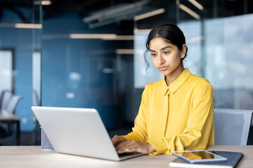 Serious young Indian businesswoman sitting at a desk in the office and working intently on a laptop