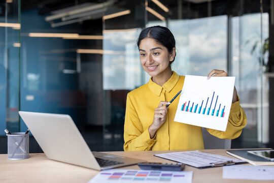 Smiling young Indian businesswoman holding a video conference, showing documents with graphs on laptop camera - Powered by Adobe
