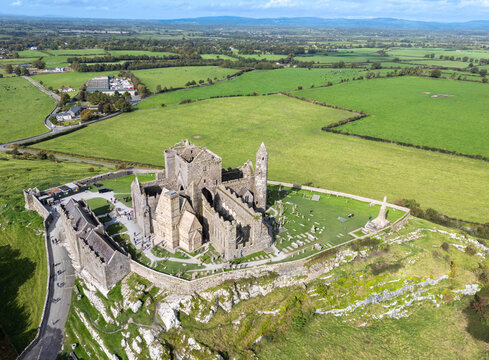 Aerial view of the Rock of Cashel, a medieval historical site atop a hill, surrounded by vibrant green fields under a vast sky, Cashel, County Tipperary, Ireland.