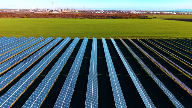 Multiple photovoltaic cells turned to the sun in the green field. Enormous oil refinery plant at backdrop. Drone footage.