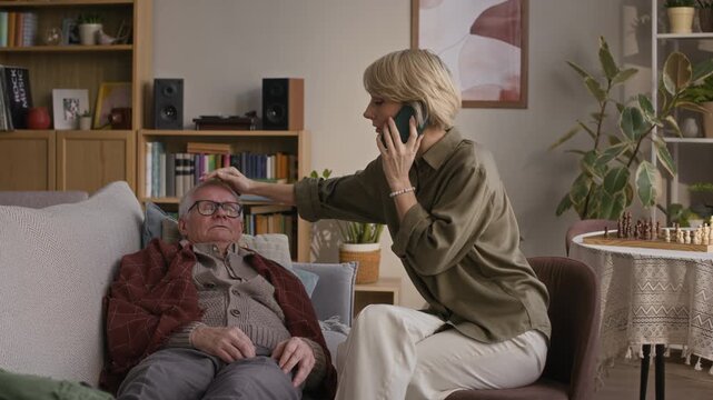 Long shot of stressed middle-aged woman calling 911 on smartphone while sitting next to senior father with fever shivering when lying on couch
