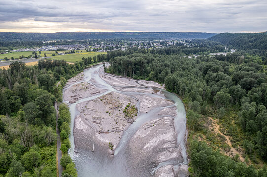 Aerial view of the winding Carbon River cutting through a landscape of dense green forests and distant buildings under a cloudy sky, Orting, Washington, United States.