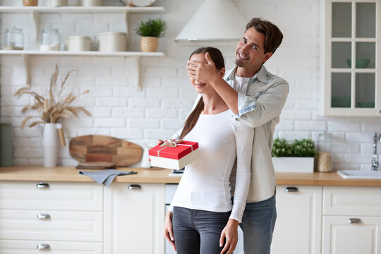 Woman in a white long-sleeve shirt holding a red gift box with a white ribbon, standing in a kitchen with a man in a light blue shirt behind her. The man is smiling and covering her eyes with his hand