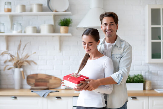 Woman in a white shirt holding a red gift box with a white ribbon, standing in a kitchen with a man in a light blue shirt behind her.
